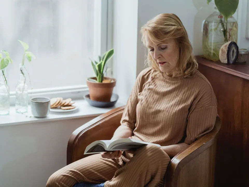 Mujer mayor leyendo un libro en un sillón junto a la ventana
