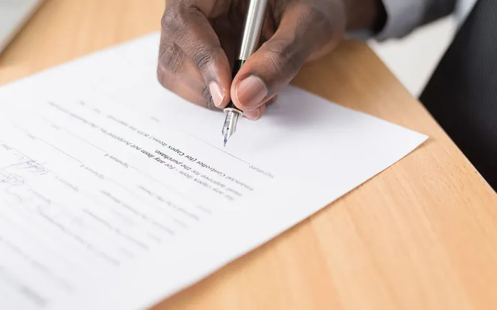 Mano firmando un documento con una pluma sobre una mesa de madera