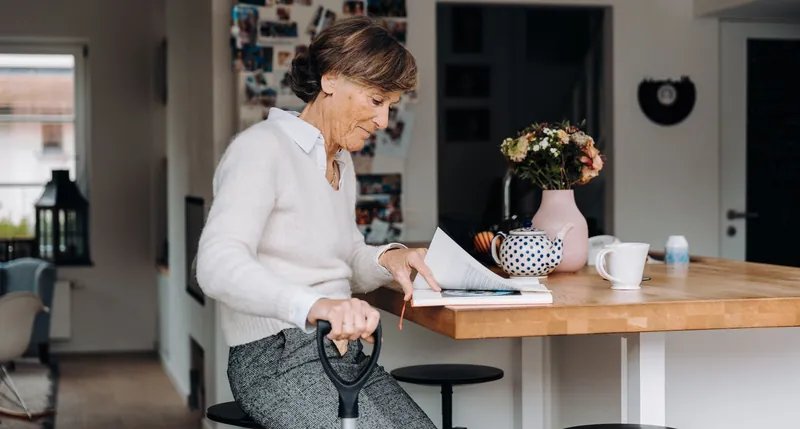 Mujer mayor leyendo una revista sentada en la barra de madera de su cocina, con tetera y flores
