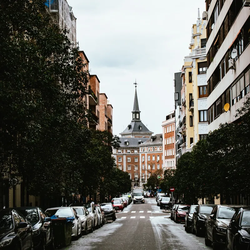 Calle residencial de Madrid con árboles, coches aparcados y una torre al fondo