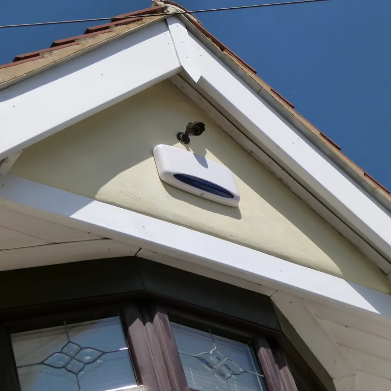 Sirena exterior de alarma blanca instalada en el hastial de una casa, junto a una cámara